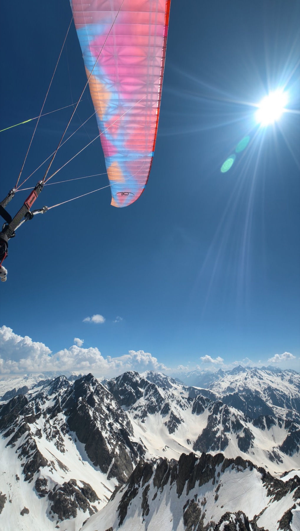 Voile rose et bleue en vol, vue de dessous, avec le soleil en contre-jour au-dessus de sommets enneigés